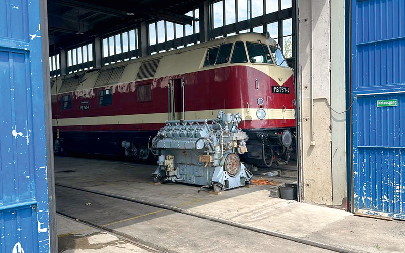 Blick in eine Halle in der eine historische Eisenbahn zu sehen ist. Davor steht ein Motor.