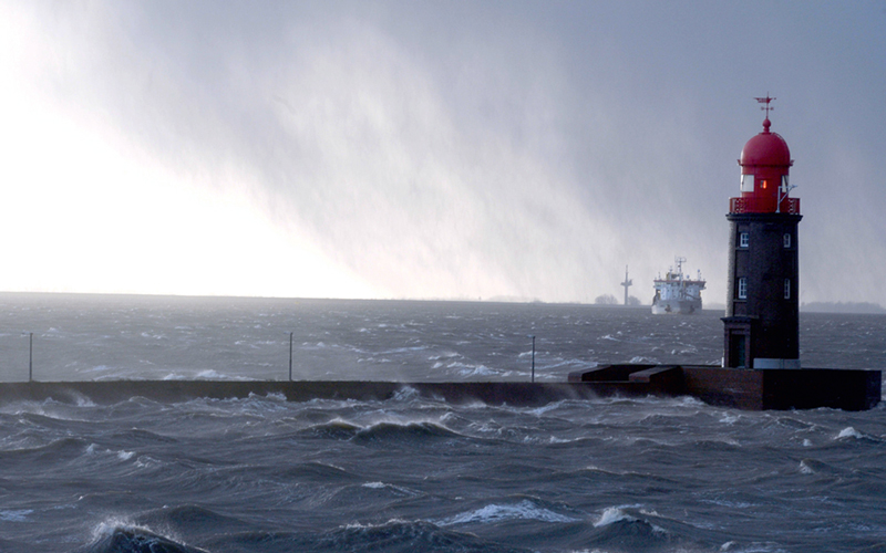 150turm8-c-bremenports_4C_800 Ein Leuchtturm mit roter Kuppel steht auf einer Mole am Meer. Im Hintergrund fährt ein großes Schiff über das stürmische Wasser. Der Himmel ist stark bewölkt, und die Atmosphäre wirkt neblig und windig – erkennbar an den bewegten Wellen und der allgemeinen Stimmung des Bildes. Die Szene vermittelt eine maritime Umgebung bei rauem Wetter.