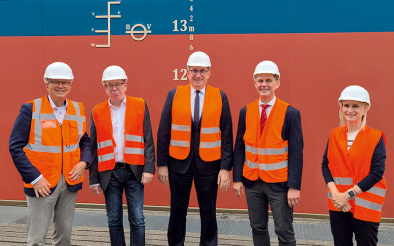 Com-01-Weilbier_Brake_c–J.MÜLLER_4C_800 Fünf Personen stehen nebeneinander vor einer großen, vertikalen Struktur – vermutlich ein Teil eines Schiffes oder einer Industrieanlage. Alle Personen tragen weiße Schutzhelme und orangefarbene Warnwesten.