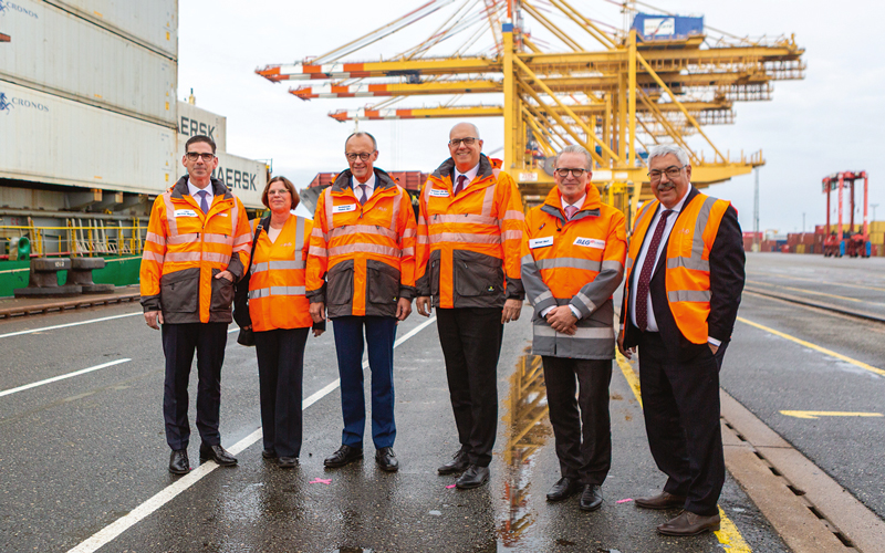 Das Foto zeigt sechs Personen in orangefarbenen Sicherheitswesten auf einem Hafengelände, im Hintergrund sind gelbe Containerkräne zu sehen. Links im Hintergrund stehen gestapelte Container. Alle tragen Anzüge bzw. formelle Kleidung, der Anlass ist ein Besuch des Bundeskanzler Friedrich Merz, der eine der Personen ist. Weiterhin sind Matthias Magnor, Kristina Vogt, Melf Grantz, Andreas Bovenschulte und Michael Blach zu sehen.