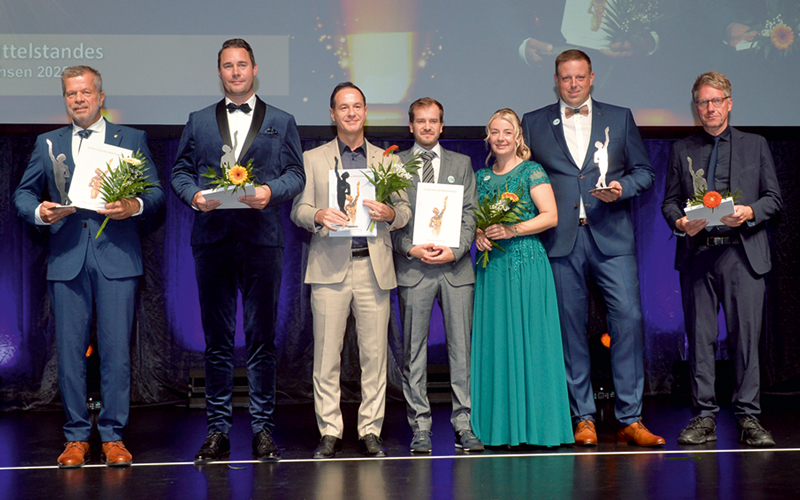 Szene bei einer Preisverleihung. Sieben Personen stehen auf einer Bühne, alle haben Blumen und Urkunden in der Hand, eine Person hält einen kleinen Pokal in der Hand.
