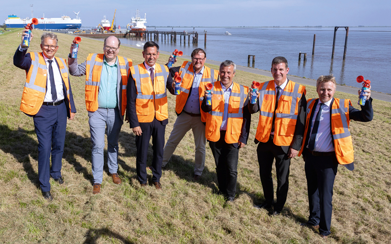 Com-02-ports-emd-rammschlag-0593-foto-andreas-burmann_4C_800 Sieben Personen mit orangenen Warnwesten stehen etwas versetzt nebeneinander, einige winken in die Kamera. Sie stehen am Strand, im Hintergrund ist Wasser zu sehen sowie ein Schiff.