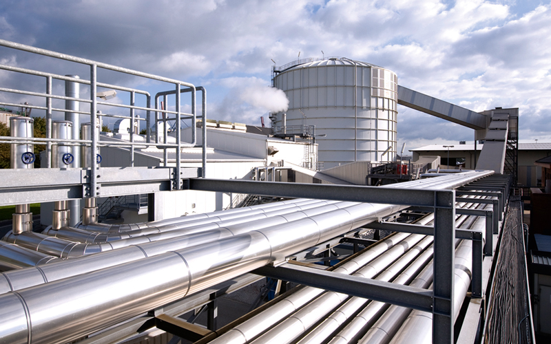 Com-03-J.MÜLLER_Schwefel—c—J.MÜLLER_4C_800 Zu sehen ist eine moderne Industrieanlage, das Schwefelterminal im Seehafen Braake. Im Vordergrund verlaufen mehrere große, horizontale Rohre, die von Metallgestellen gestützt werden. Im Hintergrund steht ein großer, zylindrischer Tank oder Silo mit einer Kuppel oben. Von diesem Tank führt ein Förderband oder eine ähnliche Struktur zu einem anderen Teil der Anlage. Der Himmel ist teilweise bewölkt.