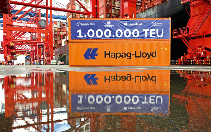 Com-03_wilhelmshaven-millionster-Container_c-Bjoern-Luebbe_4C_800 Das Bild zeigt ein Container-Terminal mit einem auffälligen orangefarbenen Container der Firma Hapag-Lloyd im Mittelpunkt. Über dem Container hängt ein großes blaues Banner mit weißer Schrift, auf dem steht: „1.000.000 TEU“. Im Hintergrund sind rote Hafenkräne und weitere Container zu sehen. Der Boden vor dem Container ist nass, wodurch dieser sich darin spiegelt, was dem Bild eine besondere visuelle Tiefe verleiht.