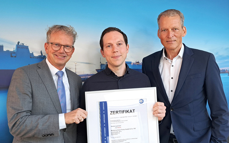 Drei Personen blicken in die Kamera, der Anlass ist die Verleihung eines Zertifikats, das die Person in der Mitte in die Kamera zeigt. Im Hintergrund ist ein großformatiges Hafenmotiv zu erkennen. Links steht der NPorts-Geschäftsführer Holger Banik, der das Zertifikat überreicht hat.