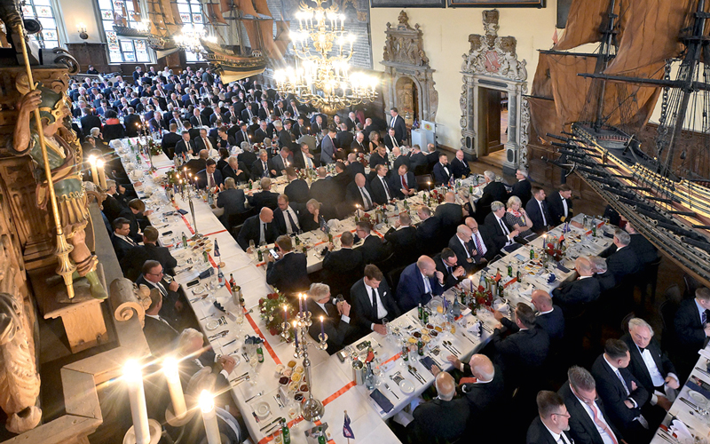 Com-10-Dinner-Kapitänstag-2025—c—BHV_4C_800 Das Foto zeigt die Obere Rathaushalle in Bremen in Vogelperspektive. Anlass ist der 58. Kapitänstag mit 300 Gästen. Die Menschen im Saal tragen dunkle Anzüge, die Mehrheit der Anwesenden sind Männer, die an eingedeckten Tischen sitzen. Man erkennt auf der rechten Seite ein großes Modell eines historischen Segelboots, die die Ausstattung im Saal ist geprägt von großen Bildern, einem riesigen Kronleuchter und alten, verzierten Fenstern.