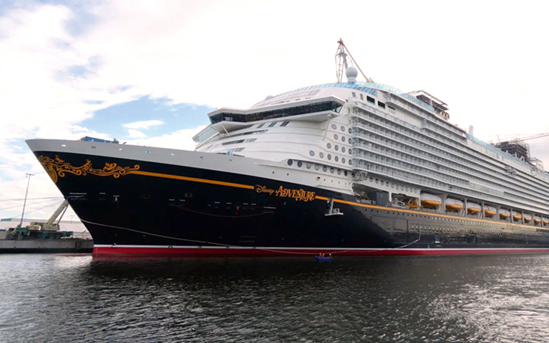 Com-14-disney-246-c-Christian-Eckert_4C_800 Zu sehen ist ein großes Kreuzfahrtschiff mit dem Namen „Disney Adventure“, das am Hafen liegt. Der Rumpf des Schiffes ist schwarz und trägt den Namen in goldenen Buchstaben. Der obere Teil des Schiffes ist weiß, mit mehreren Reihen von Fenstern und Balkonen, die zu den Kabinen gehören. An der Vorderseite sind goldene Verzierungen sichtbar, die dem Schiff ein elegantes Aussehen verleihen.