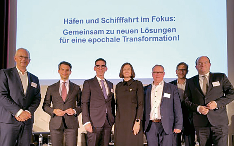 Com-17-Finanzierung-Hafeninstrastruktur_c-DVF_4C_800 Das Bild zeigt sieben Personen, die nebeneinander vor einer großen Leinwand stehen. Alle Personen tragen formelle Kleidung, wie Anzüge und Kostüme. Auf der Leinwand hinter ihnen steht in großer Schrift: „Häfen und Schifffahrt im Fokus: Gemeinsam zu neuen Lösungen für eine epochale Transformation!“ Es handelt sich um eine Veranstaltung im maritimen Bereich. Der Hintergrund ist schlicht, der Fokus liegt auf den Personen und dem Text.