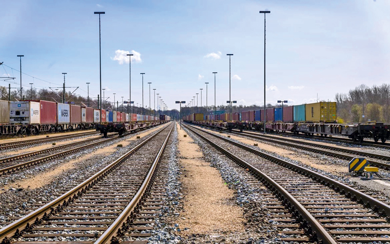 Das Bild zeigt einen großen Güterbahnhof bei Tag. Mehrere parallele Eisenbahngleise verlaufen in die Tiefe des Bildes. Auf beiden Seiten der Gleise befinden sich zahlreiche Container auf Güterwagen, die in verschiedenen Farben lackiert und mit unterschiedlichen Firmenlogos versehen sind. Entlang der Gleise stehen hohe Lampenmasten.