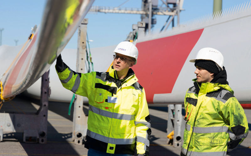 Zwei Personen mit Helmen und gelben Sicherheitsjacken an einer Projektladung bestehend aus Rotorblättern einer Windkraftanlage