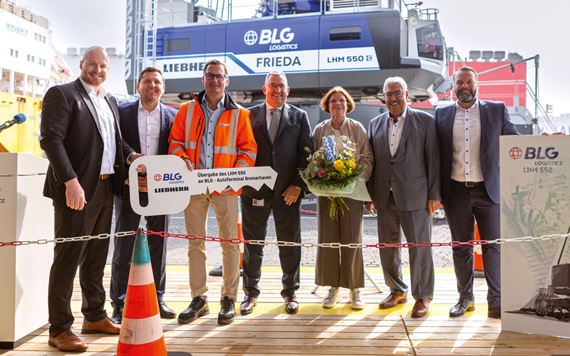 Com-22_BLG_Krantaufe_FRIEDA-3_c-BLG_4C_800 Das Bild zeigt die feierliche Übergabe eines Hafenkrans vom Typ Liebherr LHM 550. Davor stehen sechs Männer und eine Frau, Kristina Vogt. Alle tragen formelle Kleidung, einer der Männer trägt eine orangefarbene Sicherheitsweste und in der Hand einen großen Schlüssel mit der Aufschrift: „Übergabe des LHM 550 an BLG AutoTerminal Bremerhaven“ sowie den Logos von BLG und Liebherr. Frau Vogt hat einen Blumenstrauß in der Hand. Im Hintergrund ist der große Hafenkran mit der Beschriftung „BLG LOGISTICS FRIEDA LHM 550“ zu sehen. Ein Teil eines Schiffs ist ebenfalls sichtbar.