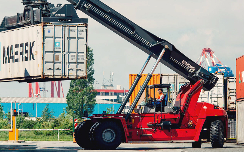 Com-23-Addicks_kreye_x_operatemybox_c-addicks-Kreye_4C_800 Zu sehen ist eine Szene aus einem Hafenbereich, in der ein großer roter Containerstapler einen weißen Container mit der Aufschrift „MAERSK“ anhebt. Rundherum sind weitere Container in verschiedenen Farben zu sehen, darunter orangefarbene und blaue. Im Hintergrund erkennt man Hafenkräne und andere Strukturen, die typisch für Umschlagplätze sind.