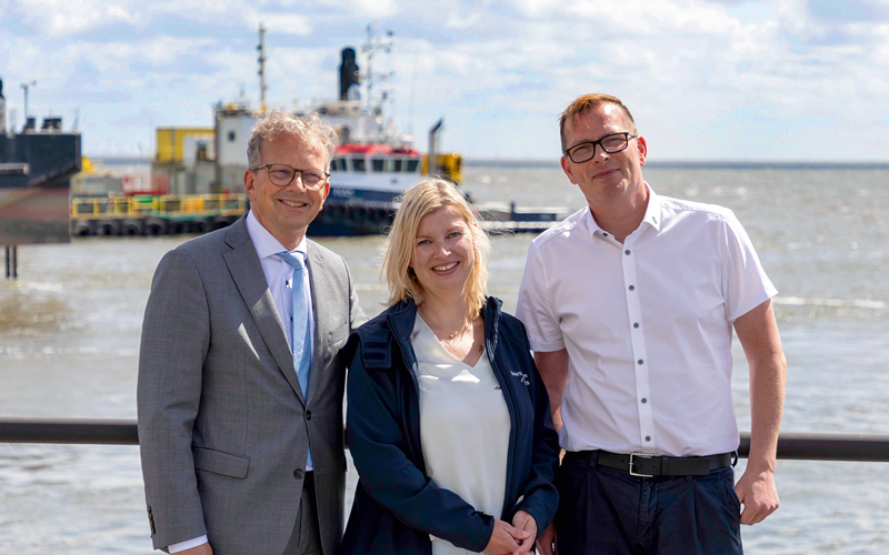 Holger Banik, Alexandra Brandt und Knut Kokkelink mit Wasser und einem Schiff im Hintergrund