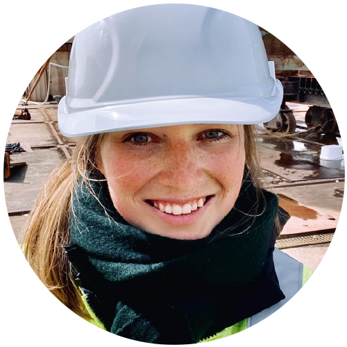 Portrait of Juliane Bellstedt, wearing a yellow safety vest and white helmet, with a ship's hull visible in the background, presumably a shipyard