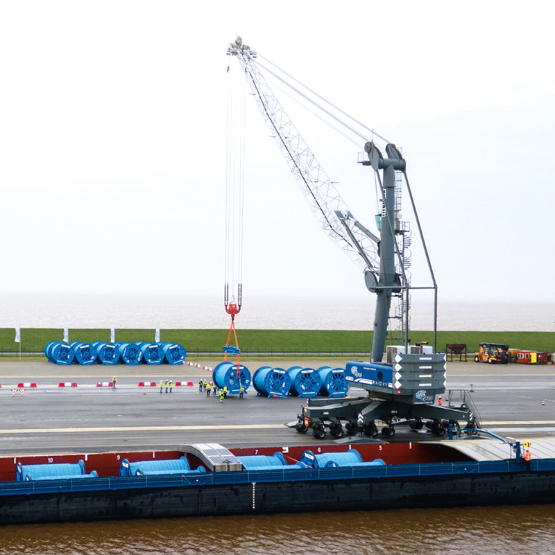 Ein mobiler Kran verlädt Kabeltrommeln auf einen kleinen Frachter im Kanal.