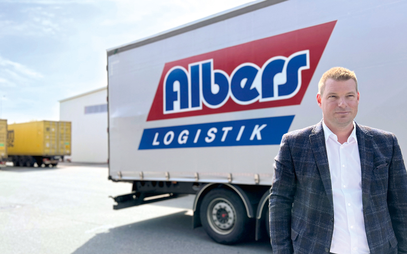 Pascal Müller vor einem Trailer auf dem das blau, weiß, rote Logo von Albers Logistik zu sehen ist. Müller trägt ein dunkles Jacket und ein weißes Hemd.