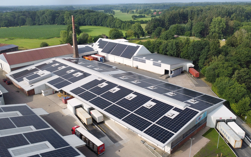 Drohnenaufnahme von Logistikhallen deren Dächer großflächig mit Photovoltaikanlagen ausgestattet sind. Vor den Hallen sind mehrere Fahrzeuge zu sehen, die Umgebung ist ländlich.