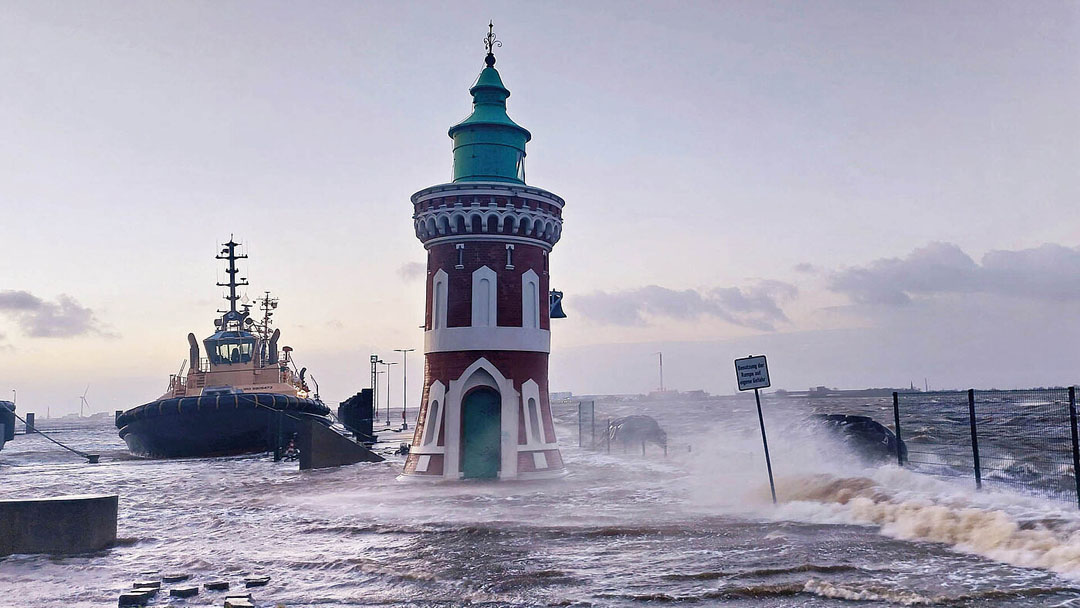 Storm surge at a port area: High waves flood a quay wall, with a historic lighthouse standing in the center. Tugboats are anchored in the background.  