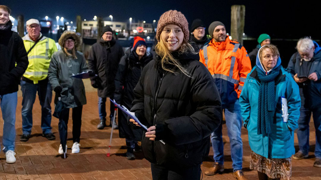 Mehrere Personen betrachten die Beleuchtung bei der Informationsveranstaltung. Es ist dunkel und im Hintergrund sind Hafenanlagen und Beleuchtung zu sehen.