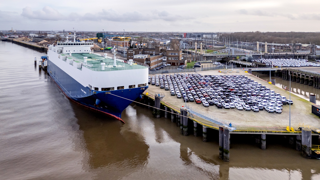 Vogelperspektive auf einen Autofrachter am Terminal, an Land stehen unzählige Autos.