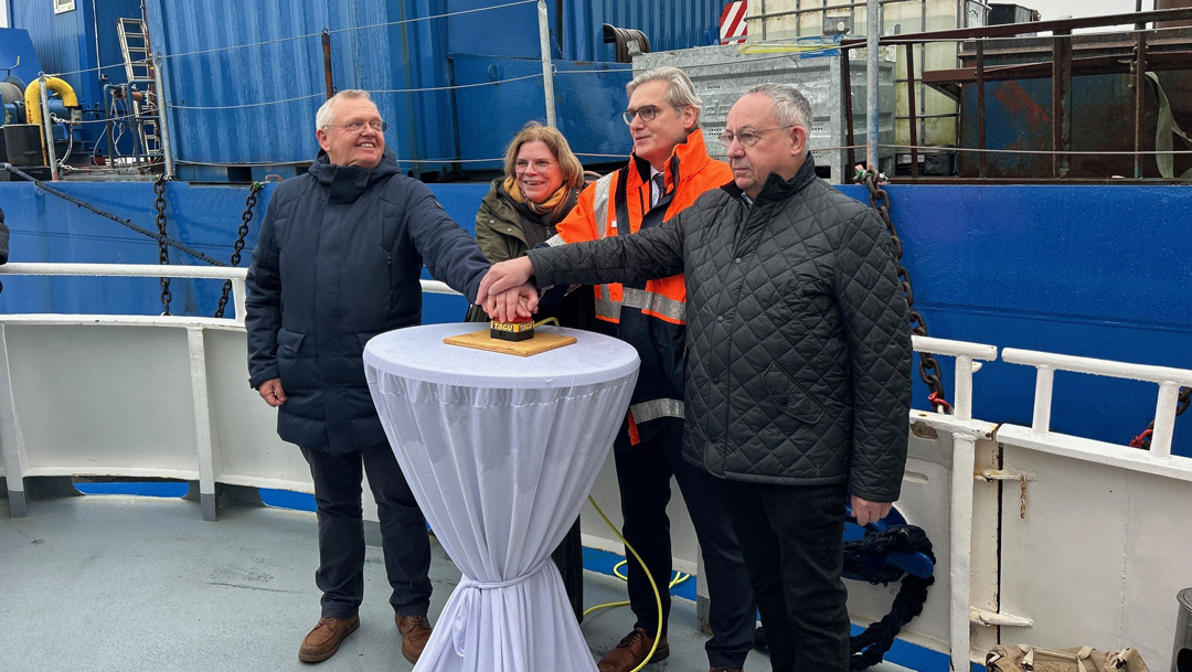 Logistics Pilot_Neubau Nordmole Bremerhaven Rammschlag Vier Personen, darunter Kristina Vogt, an einem Stehtisch auf dem sie gleichzeitig eine Art Buzzer drücken. Das Setting ist maritim.