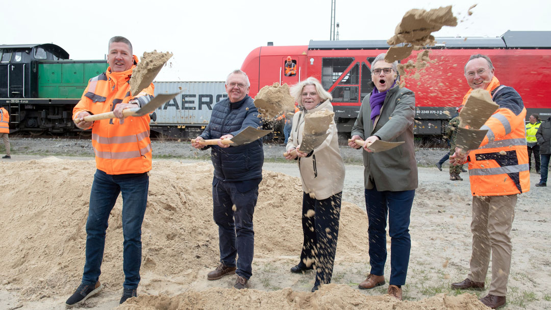 Fünf Personen haben einen Spaten in den Händen und werfen Sand hoch, im Hintergrund sind Waggons und Züge zu sehen