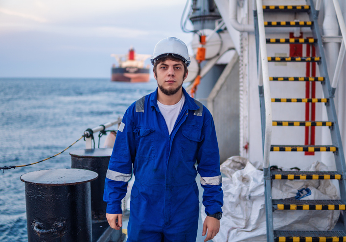 Junger Mann an Deck eines Offshore-Schiffes, mit Helm und Arbeitsoverall.
