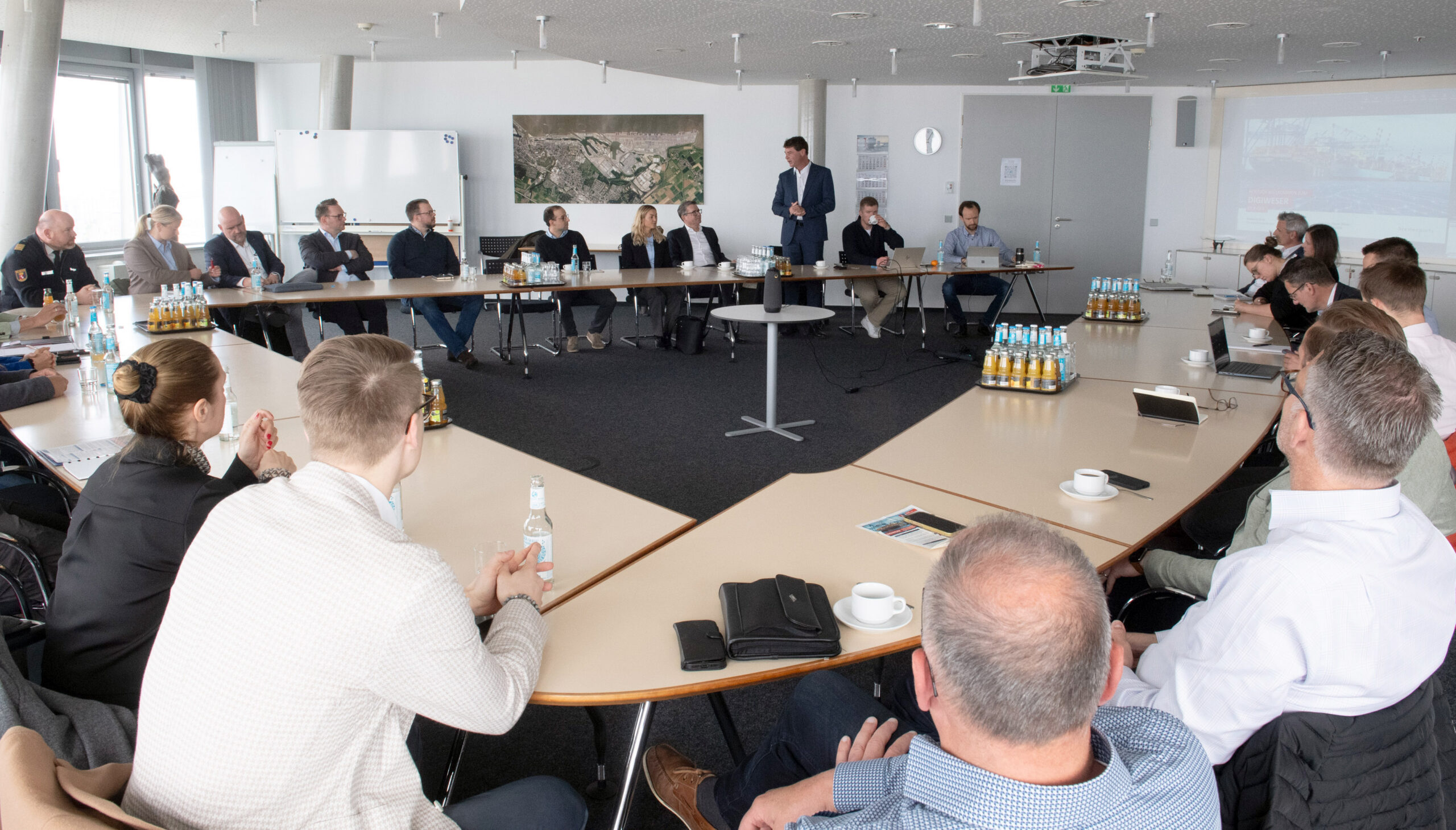 Blick in einen Konferenzraum in dem rund 25 Personen an einem runden Tisch sitzen. Eine Person steht und spricht zu den restlichen Anwesenden.