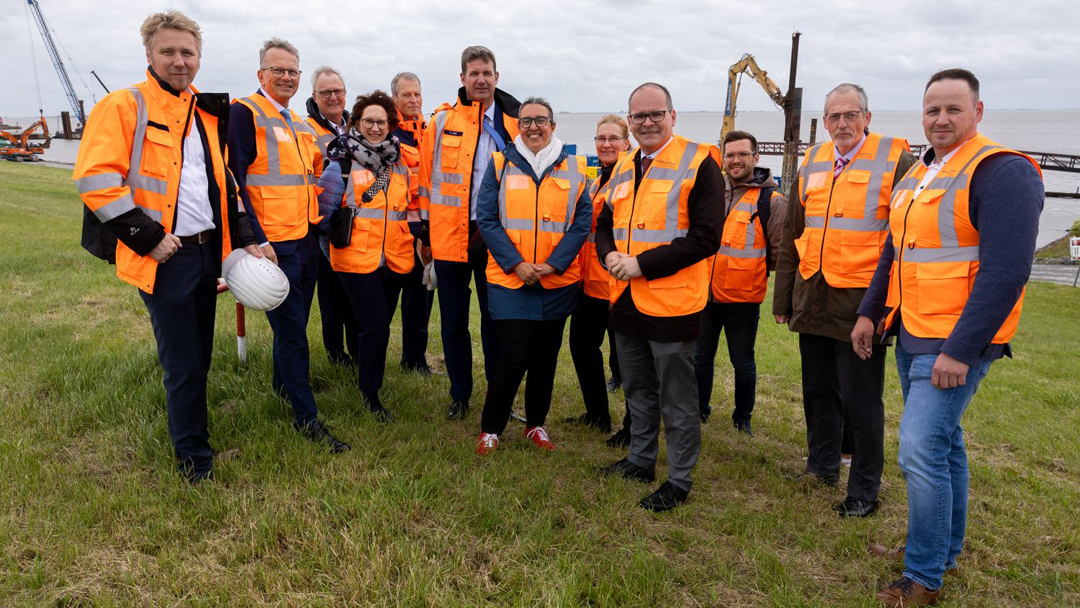 Baustellenbehung in Emden gruppenbild von zwölf Personen, alle mit orangenen Warnwesten. Im Hintergrund ist ein Baustellenfahrzeug sowie Wasser zu sehen.