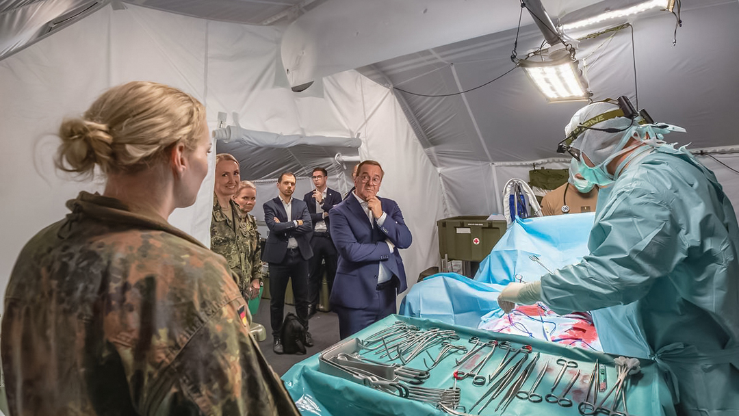 Boris Pistorius bei einem Besuch der Bundeswehr Der Verteidigungsminister steht in einem Sanitätszelt und hört aufmerksam einer Mitarbeiterin der Bundeswehr zu. Im Vordergrund sieht man einen Arzt an einem OP Tisch. Neben Herrn Pistorius stehen noch zwei weitere Herren im Anzug sowie zwei Mitarbeiterinnen der Bundeswehr.
