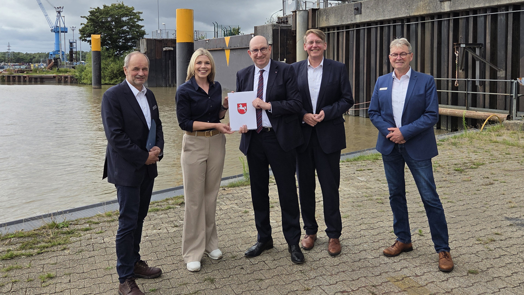 Übergabe eine Förderbescheids für Stadt Papenburg Gruppenfoto mit fünf Personen. Eine Person bekommt ein Dokument übergeben mit dem niedersächsischen Logo auf der Vorderseite, das deutlich in die Kamera gehalten wird. Im Hintergrund ist eine Schleusenanlage zu sehen.