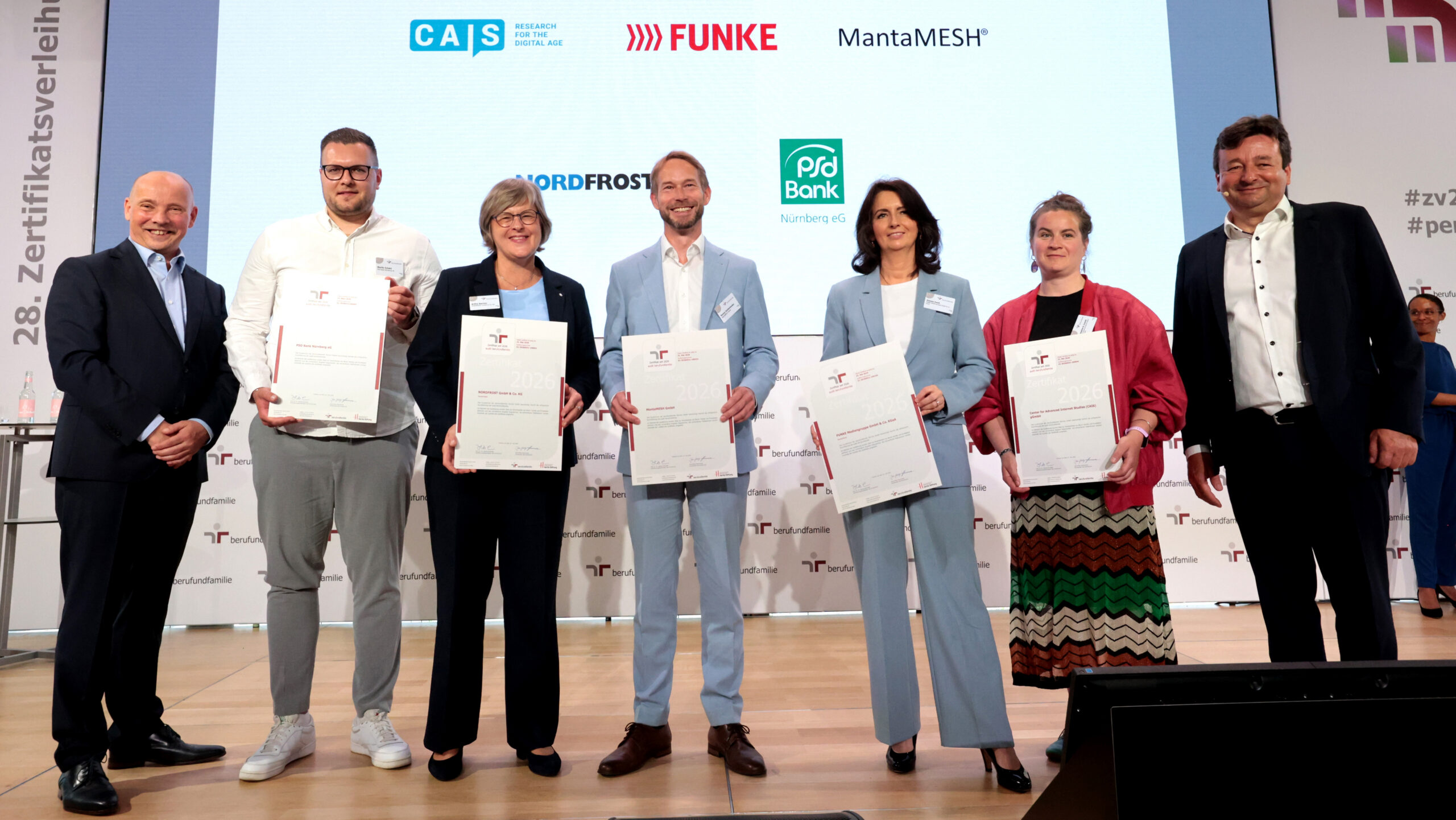 Seven people are standing on a stage in front of a wall displaying sponsors’ logos. Five of them are holding certificates up to the camera.