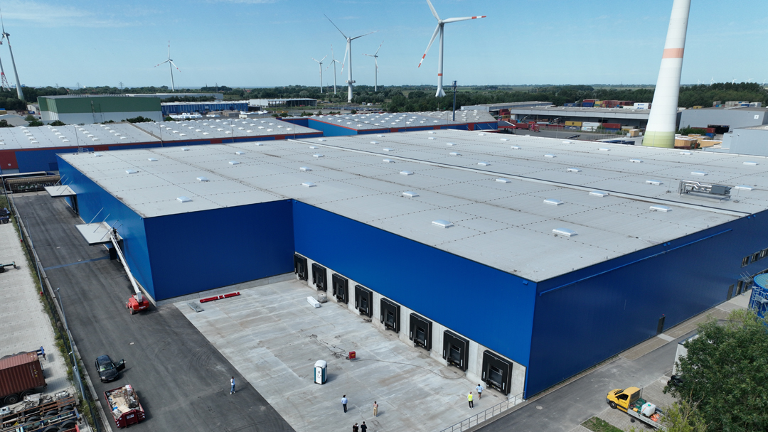 Luftaufnahme großer blauer Logistikhallen, im Hintergrund stehen mehrere Windräder