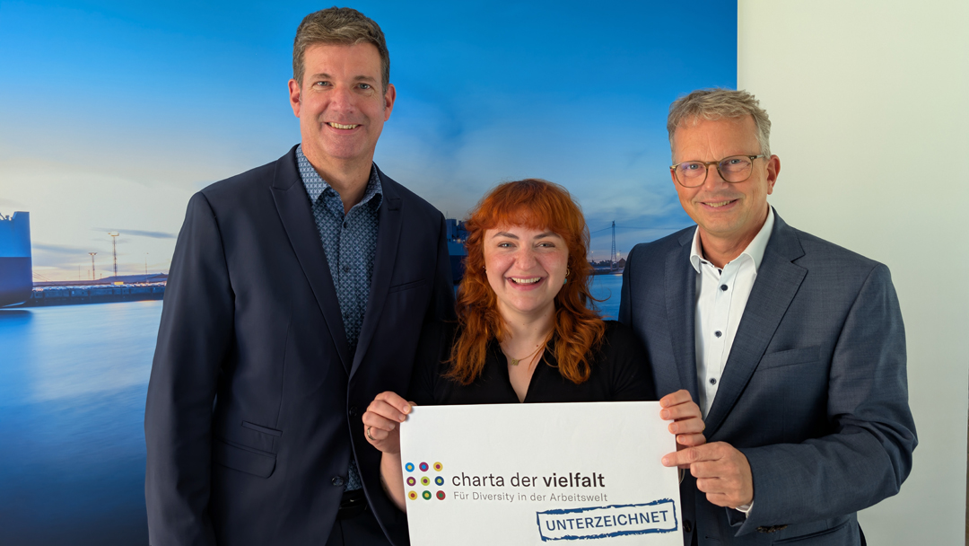 Drei Personen, zwei Männer und in der Mitte eine Frau, blicken lächelnd in die Kamera und halten symbolisch ein Plakat hoch auf dem in bunten Buchstaben steht "Charta der Vielfalt" und darunter als Stmpelschrift "Unterzeichnet"