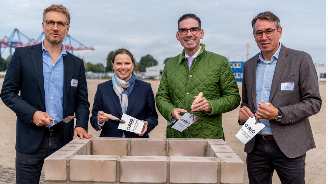Grundsteinsteinlegung_ BLG LOGISTCS Vier Personen die vor einer kleinen frisch gemauerten Grundmauer stehen. In den Händen halten sie symbolische Maurerkellen mit BLG Aufdruck. Im Hintergrund sind Hafenkräne erkennbar.