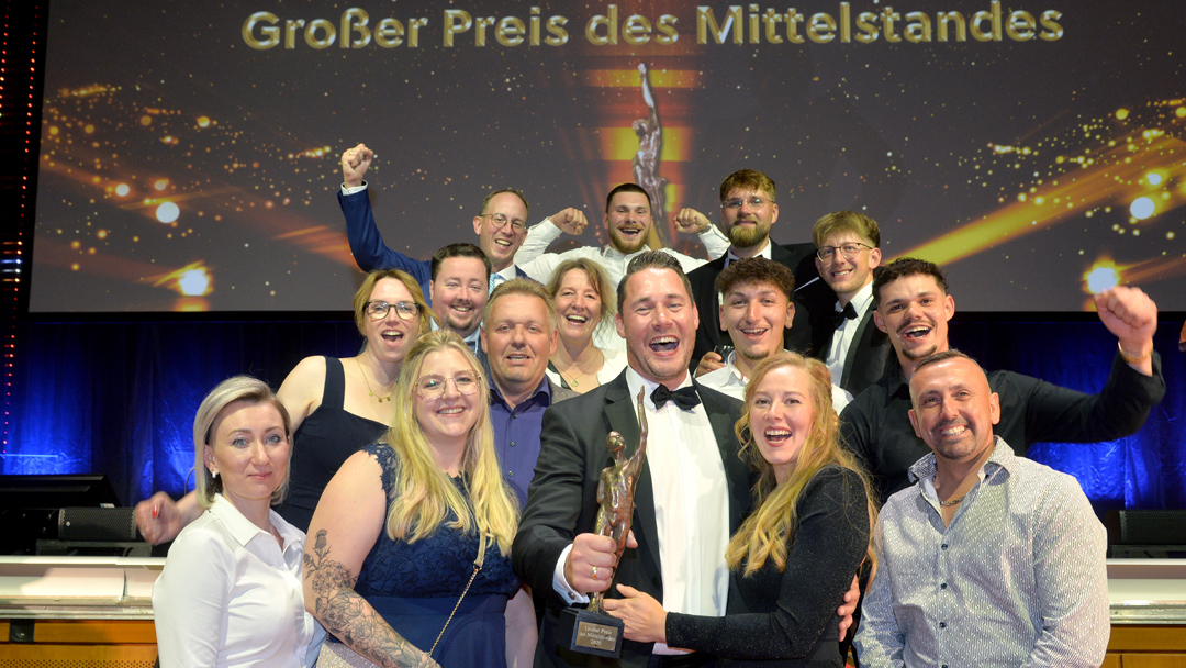 Gruppenfoto von 15 festlich gekleideten Personen, die freudig lachend in die KAmera schauen und teilweise die Arme jubelnd in die Höhe strecken. Eine Person in der ersten Reihe hält einen Preis in Form einer kleinen Skulptur in der Hand. Im Hintergrund steht auf einer Leinwand "Großer Preis des MIttelstands"
