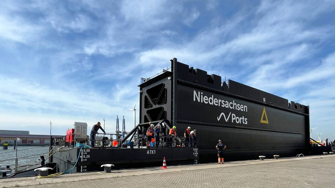 Ein riesiges schwarzes Schleusentor mit Niedersachsen Ports-Schriftzug wird zu Wasser gelassen. Mehrere Mitarbeiter sind daran beteiligt.