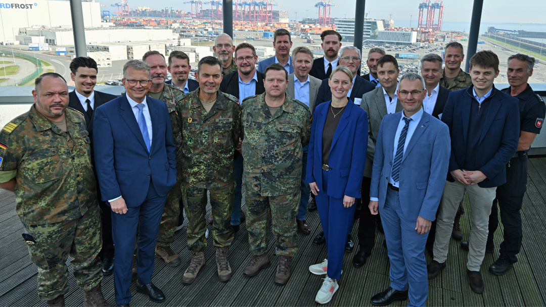 Austausch JadeWeserPort mit Bundeswehr GRuppenfoto von rund 20 Personen, davon eine Frau. Fünf Personen tragen Bundeswehrkleidung, die anderen Anzüge. Eine PErson in Polizeiuniform ist zu sehen. Im Hintergrund sind Hafenanlagen zu sehen.
