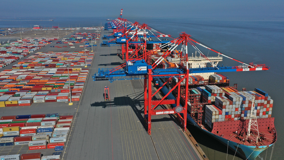 Blick auf ein Containerterminal. Rote Kräne löschen ein Containerschiff, das rechts am Kai anlegt. Auf der linken Seite sind reihenweise bunter Container zu sehen.